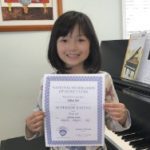 Here's an alt tag for the image: Smiling girl holding piano achievement certificate.